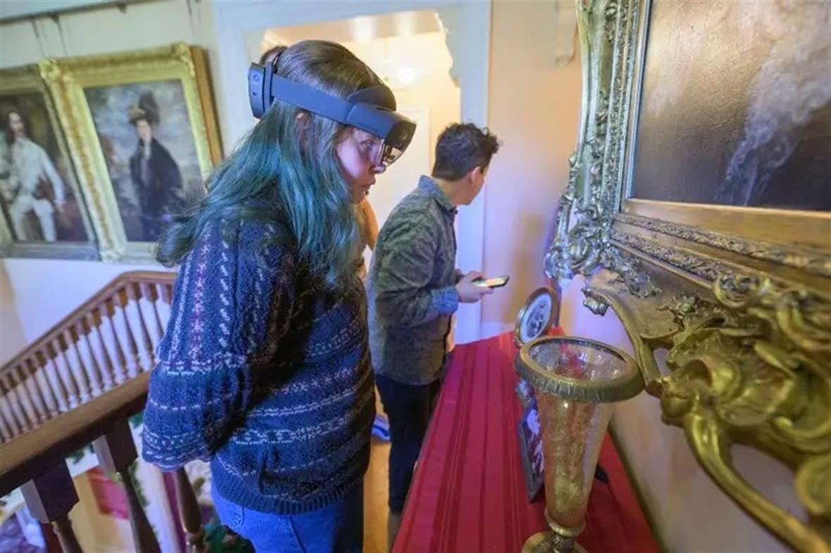 A student wearing pattered sweater and augmented reality googles looks at an art piece while another student observes another piece in the background