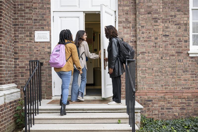 Three female students in coats and backpacks enter UD's Student Success Center through the white door of a historic red brick building.